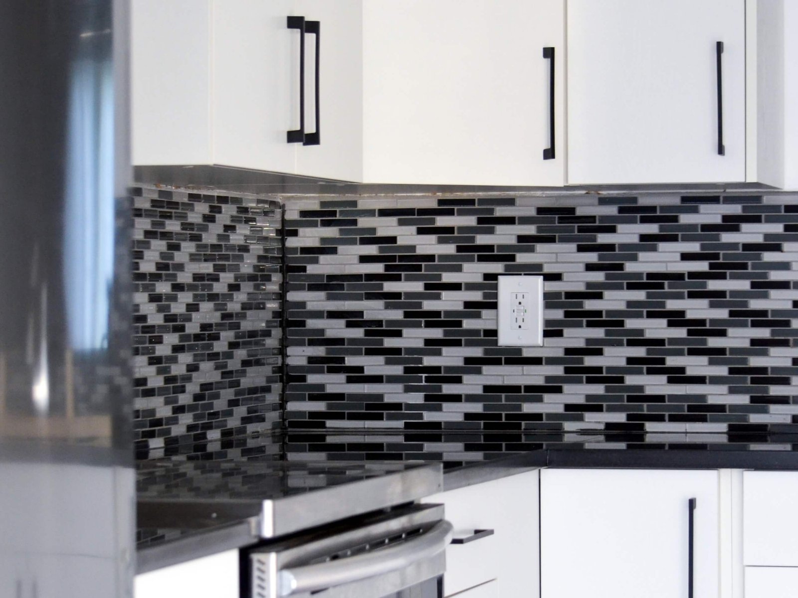 A close-up of a modern kitchen featuring a black, grey, and silver glass mosaic tile backsplash paired with white cabinets and matte black handles. The reflection from the stainless steel stove and the sleek countertop highlights the contemporary, high-contrast design.