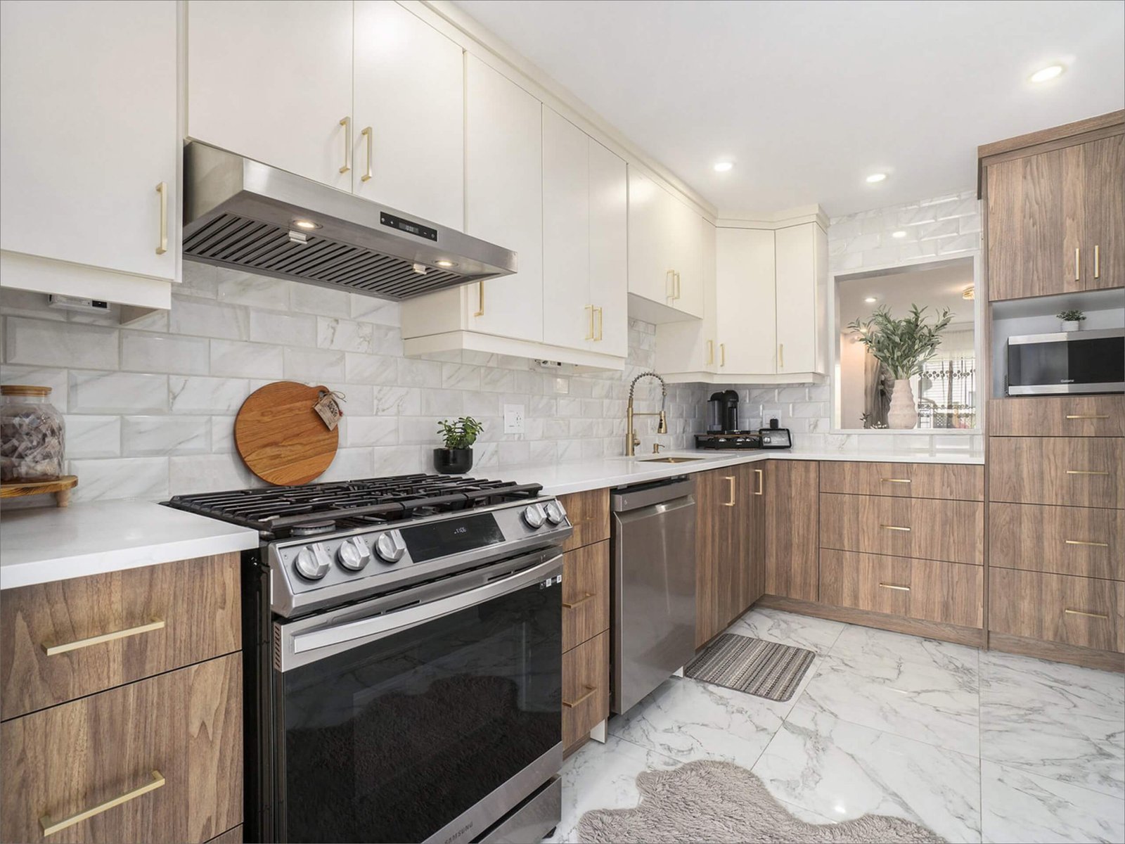 A bright modern kitchen featuring white upper cabinets, warm wood-grain lower cabinets with gold handles, and polished white quartz countertops. A stainless steel gas range, dishwasher, and hood vent sit against a glossy marble-patterned backsplash. Large marble tile flooring reflects the natural light, while a small plant, cutting board, and soft rug add warmth to the space.