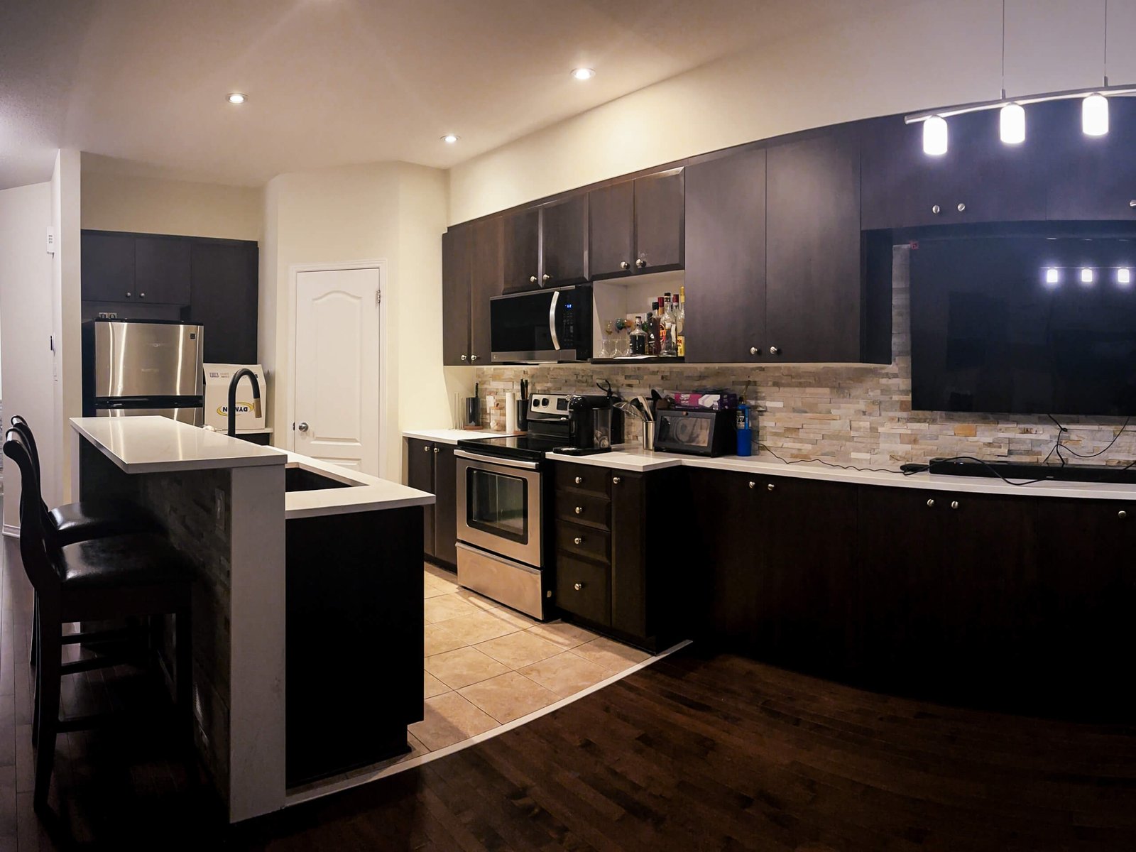 A modern open-concept kitchen with dark espresso cabinets, white quartz countertops, stainless steel appliances, and warm recessed lighting. A raised peninsula with bar stools sits on the left, while a long counter with a stone backsplash runs along the right wall beneath a mounted TV and pendant lights. The space features a mix of tiled and hardwood flooring, creating a cozy, contemporary look.