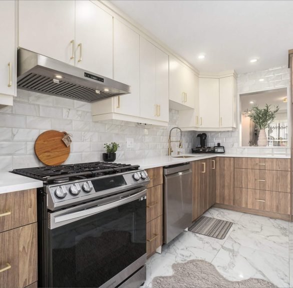 A bright modern kitchen featuring white upper cabinets, warm wood-grain lower cabinets with gold handles, and polished white quartz countertops. A stainless steel gas range, dishwasher, and hood vent sit against a glossy marble-patterned backsplash. Large marble tile flooring reflects the natural light, while a small plant, cutting board, and soft rug add warmth to the space.