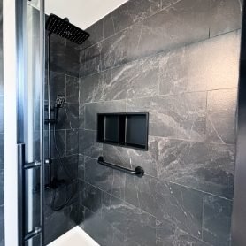A modern walk-in shower featuring large dark grey stone-look tiles, a matte black rainfall showerhead, matching handheld spray, a built-in black niche shelf, and a sleek black grab bar.