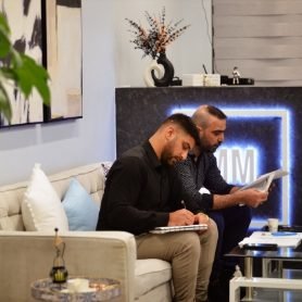 Mahmoud with his brother sitting on a sofa in a modern office lounge, reviewing paperwork and taking notes. Behind them is a glowing blue MM Brothers sign, decorative artwork, and a vase arrangement on a shelf.