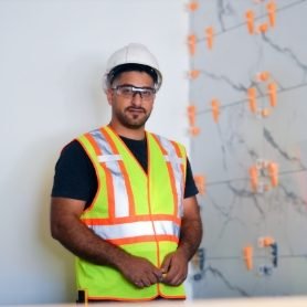 Mahmoud Allush wearing a hard hat, safety glasses, and a reflective safety vest stands in front of a tiled wall installation with orange spacers, posing confidently on site.